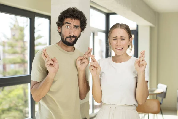 young adult couple crossing fingers anxiously and hoping for good luck with a worried look