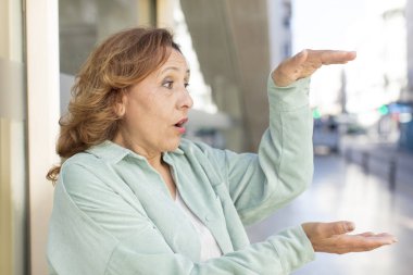 Orta yaşlı bir kadın bir objeyi iki eliyle fotokopi alanında tutuyor, bir objeyi gösteriyor, sunuyor ya da reklamını yapıyor.