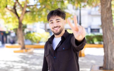 Genç yakışıklı adam gülümsüyor ve mutlu görünüyor, kaygısız ve olumlu, zafer veya barış işareti yapıyor.