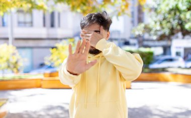 Genç ve yakışıklı bir adam elleriyle yüzünü kapatıyor ve kamerayı durdurmak için diğer elini öne koyuyor. Fotoğraf ya da resimleri reddediyor.