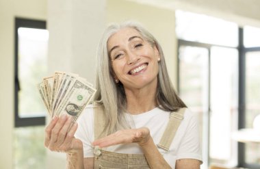 Güzel, son sınıf öğrencisi, neşeli bir şekilde gülümseyen, mutlu hisseden ve bir konsept gösteren bir kadın. Dolar banknotlarıyla