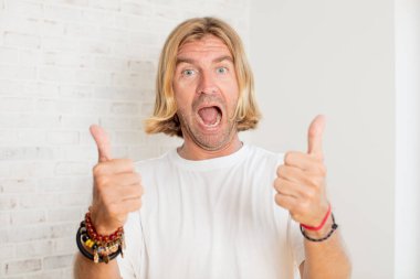 Genç, sarışın, yetişkin bir adam neşeyle gülümsüyor ve mutlu görünüyor. Kaygısız ve pozitif hissediyor. İki başparmağı da havada.