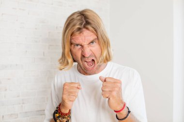 Genç, sarışın, yetişkin bir adam sinirli, sinirli, sinirli bakışlar ve sıkı yumruklar ile agresif bir şekilde bağırıyor.
