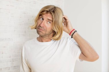 Genç sarışın yetişkin bir adam kafası karışmış ve kafası karışmış hissediyor, başını kaşıyor ve yan tarafa bakıyor.