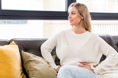 Genç, güzel bir kadın mutlu, neşeli ve kendinden emin görünüyor. Gururla gülümsüyor ve iki elini de kanepede oturan kalçalarına koyuyor.