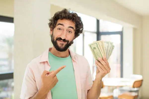 crazy bearded man smiling cheerfully, feeling happy and pointing to the side. dollar banknotes concept