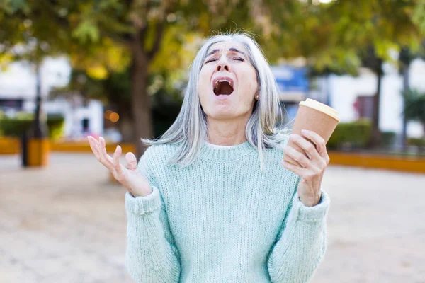 pretty senior woman screaming with hands up in the air with a take away coffee