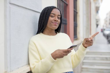 Siyah afro kadın neşeyle gülümsüyor, mutlu hissediyor ve bir tarafı işaret ediyor. akıllı telefon konseptini kullanarak