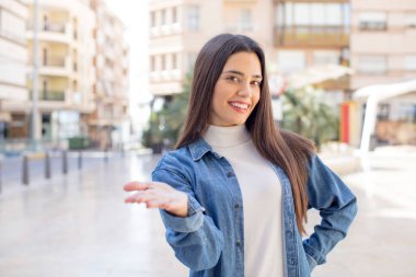 Güzel, genç, yetişkin bir kadın gülümsüyor, mutlu, kendinden emin ve arkadaş canlısı görünüyor, bir anlaşmaya varmak için el sıkışmayı teklif ediyor, işbirliği yapıyor.