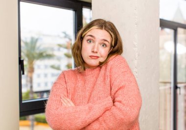 Güzel, beyaz bir kadın omuz silkiyor, kafası karışık ve kararsız, kolları bağlı ve şaşkın görünüyor. ev içi konsept