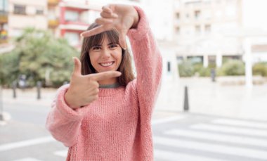 Güzel genç bir kadın mutlu, arkadaş canlısı ve pozitif hissediyor, gülümsüyor ve elleriyle bir portre veya fotoğraf çerçevesi yapıyor.