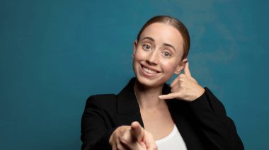 Neşeli bir şekilde gülümsüyor ve telefonla konuşurken kamerayı işaret ediyor.