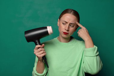 Yeşil stüdyo arka planında saç kurutma makinesi olan genç sarışın kadın.
