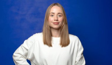 Mutlu bir şekilde gülümsüyor, eli kalçasında ve kendinden emin, olumlu, gururlu ve arkadaş canlısı bir tavırla.