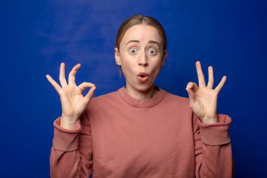 Sarışın kadın şok olmuş, şaşırmış ve şaşırmış hissediyor. Onaylıyor. İki eliyle de onay işareti yapıyor.