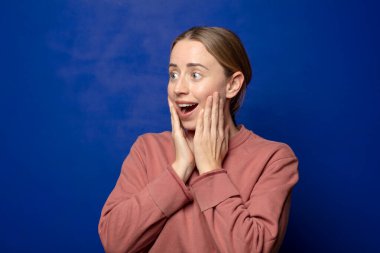 Sarışın kadın mutlu, heyecanlı ve şaşırmış hissediyor, iki eli yüzü yana bakıyor.