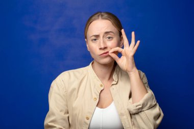 Sarışın kadın ciddi ve hoşnutsuz görünüyor. İki parmağın önde olsun, reddedilmek üzere, sessizlik istiyor.