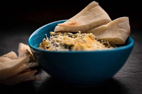 Teal colored bowl of hot spinach and artichoke dip with pieces of toasted pita bread around.