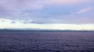 Ocean coastline with wind power turbines spread on the shore during stormy, cloudy day with blue cast as seen from a ship sailing along them