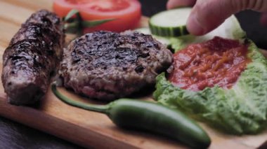 Barbecued ground meat patty and sausage on wooden surface garnished with veggies