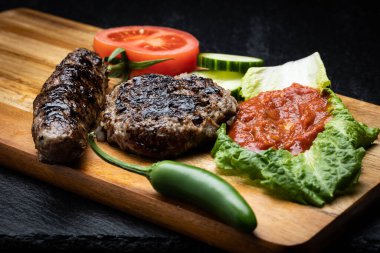Grilled sausage and a patty with vegetables and spices on wooden background