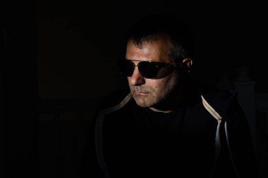 Siyah arka planda, orta yaşlı, güneş gözlüklü yakışıklı bir adamın portresi.