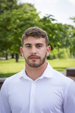 Sakallı yakışıklı bir gencin portresi, parkta duruyor ve eğleniyor..