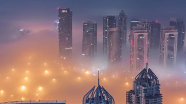 Şeyh Zayed Yolu yakınlarındaki JLT gökdelenleri gece gündüz geçiş zamanı. Aydınlanmış konut binaları ve kavşaktaki trafik. Güneş doğmadan önce sisli bir sabah