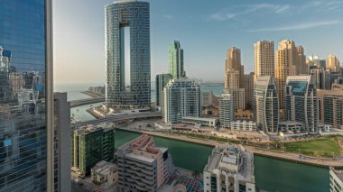Panorama, Dubai Marina gökdelenlerini ve JBR bölgesini lüks binalar ve tatil köylerinin zaman çizelgeleriyle gösteriyor. Palmiyeler ve kanalda yüzen teknelerle dolu bir liman.