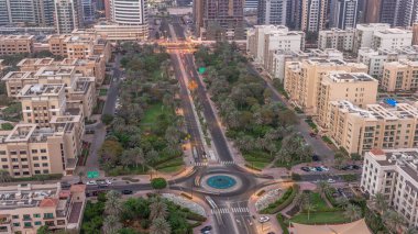 Yeşiller Bölgesi hava sahasındaki düşük katlı binalar gece ve gündüz geçiş zamanları. Palmiyeler ve ağaçlarla dolu Dubai silueti. Bir yol kavşağında trafik