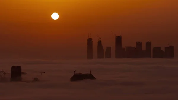 高層ビルや塔が朝の霧の空中のタイムラプスで覆われているドバイクリークハーバーの日の出 ビルやオレンジの空の後ろに大きな赤い太陽が昇る — ストック写真