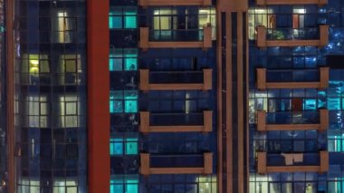 Aydınlatılmış çalışma odası saatleriyle apartman penceresi ön cephesinin gece görüşü. Balkonlu gökdelende parlayan ışık