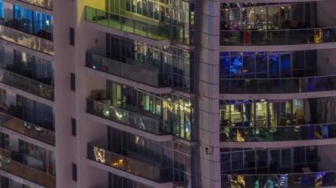 Dışarıdaki apartmanın gece manzarası renkli zaman çizelgesi. Pencerelerinde parlayan ışıklar olan yüksek katlı bir gökdelen. Balkonda sandalyeler
