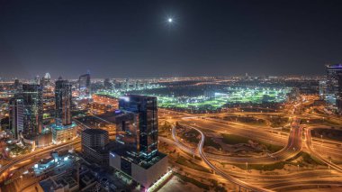 Panorama showing media city, Dubai marina and JLT illuminated skyscrapers along Sheikh Zayed Road with big crossroad junction aerial night . Rising moon over residential and office buildings and golf course from above.