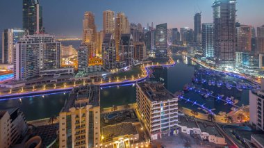 Dubai Marina with boats and yachts parked in harbor and illuminated skyscrapers around canal aerial night to day transition  before sunrise. Towers of JBR district on a background