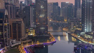 Dubai Marina with boats and yachts parked in harbor and illuminated skyscrapers around canal aerial night to day transition  before sunrise. Towers along waterfront