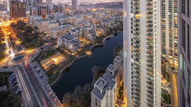 Pond and low rise buildings in Greens district aerial day to night transition . Dubai skyline with skyscrapers in Barsha Heights district on a background