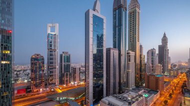 Aerial view of Dubai International Financial District with many skyscrapers night to day transition . Traffic on a road near metro station and multi storey parking with rooftop swimming pool. Dubai, UAE.