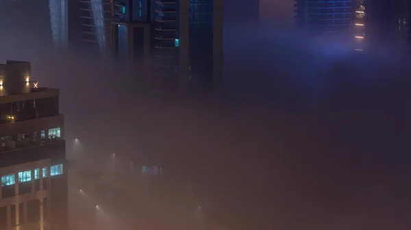 Buildings are covered in thick layer of fog in Business Bay night . Illuminated skyscrapers around water canal with traffic on a road aerial top view