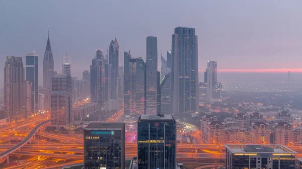 Panorama of Dubai Financial Center district with tall skyscrapers with illumination night to day transition . Aerial view to towers with morning fog before sunrise