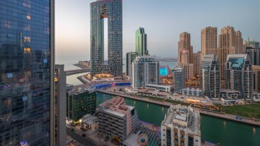 Günbatımından sonra Dubai Marina gökdelenleri ve JBR semti aydınlanmış lüks binalar ve gece geçiş zamanlarıyla dolu tatil köyleri. Palmiyeler ve kanalda yüzen teknelerle dolu bir liman.