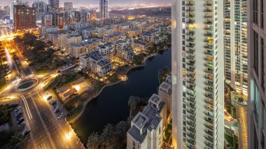 Yeşiller bölgesindeki havuz ve alçak katlı binalar gündüz ve gece geçiş zamanları. Arka planda Barsha Heights bölgesinde gökdelenleri olan Dubai gökdelenleri