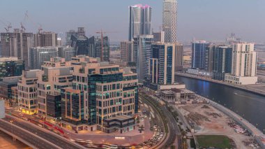 Körfez Meydanı 'nda günden geceye geçiş. Karışık kullanım ve Dubai 'deki Business Bay' de bulunan düşük katlı kompleks ofis binaları. Yukarıdan hava manzaralı ve yolda trafik var.