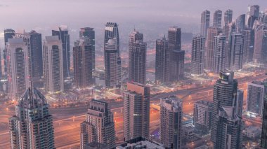JLT skyscrapers and marina towers near Sheikh Zayed Road aerial night to day transition . Illuminated residential buildings and skyline with villas. Foggy morning before sunrise