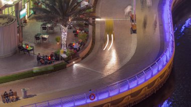 Dubai Marina hava sahasında palmiyelerle rıhtım gezintisi. İnsanlar bir bankta yürüyor ve oturuyor. Yatlar ve tekneler suda yüzüyor.