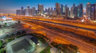 Dubai Marina gökdelenleri ve Şeyh Zayed yolu ile metro demiryolu havadan geceye geçiş panoramik. Günbatımından sonra modern kulelerin yakınındaki bir otoyolda trafik, Birleşik Arap Emirlikleri