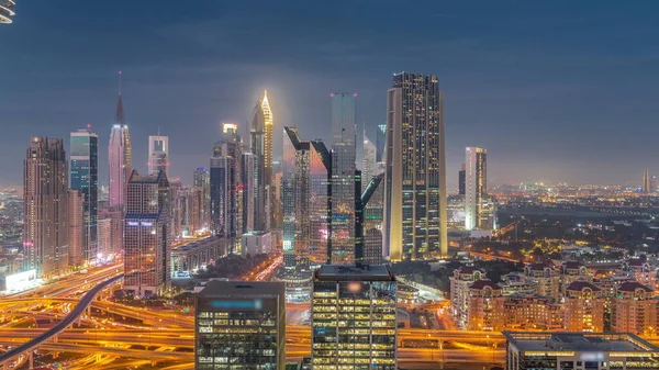 Panorama of Dubai Financial Center district with tall skyscrapers with illumination day to night transition . Aerial view to towers along busy highway after sunset