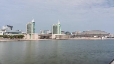 Modern tall residential and office buildings reflecting in the water under blue cloudy sky timelapse hyperlapse in Lisbon, Portugal. Park nations luxury district with concert hall and flags