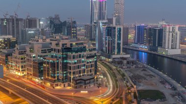 Körfez Meydanı 'nda günden geceye geçiş. Karışık kullanım ve Dubai 'deki Business Bay' de bulunan düşük katlı kompleks ofis binaları. Yukarıdan hava manzaralı ve yolda trafik var.