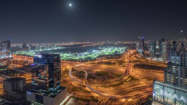 Panorama showing media city, Dubai marina and JLT illuminated skyscrapers along Sheikh Zayed Road with big crossroad junction aerial night. Rising moon over residential and office buildings and golf